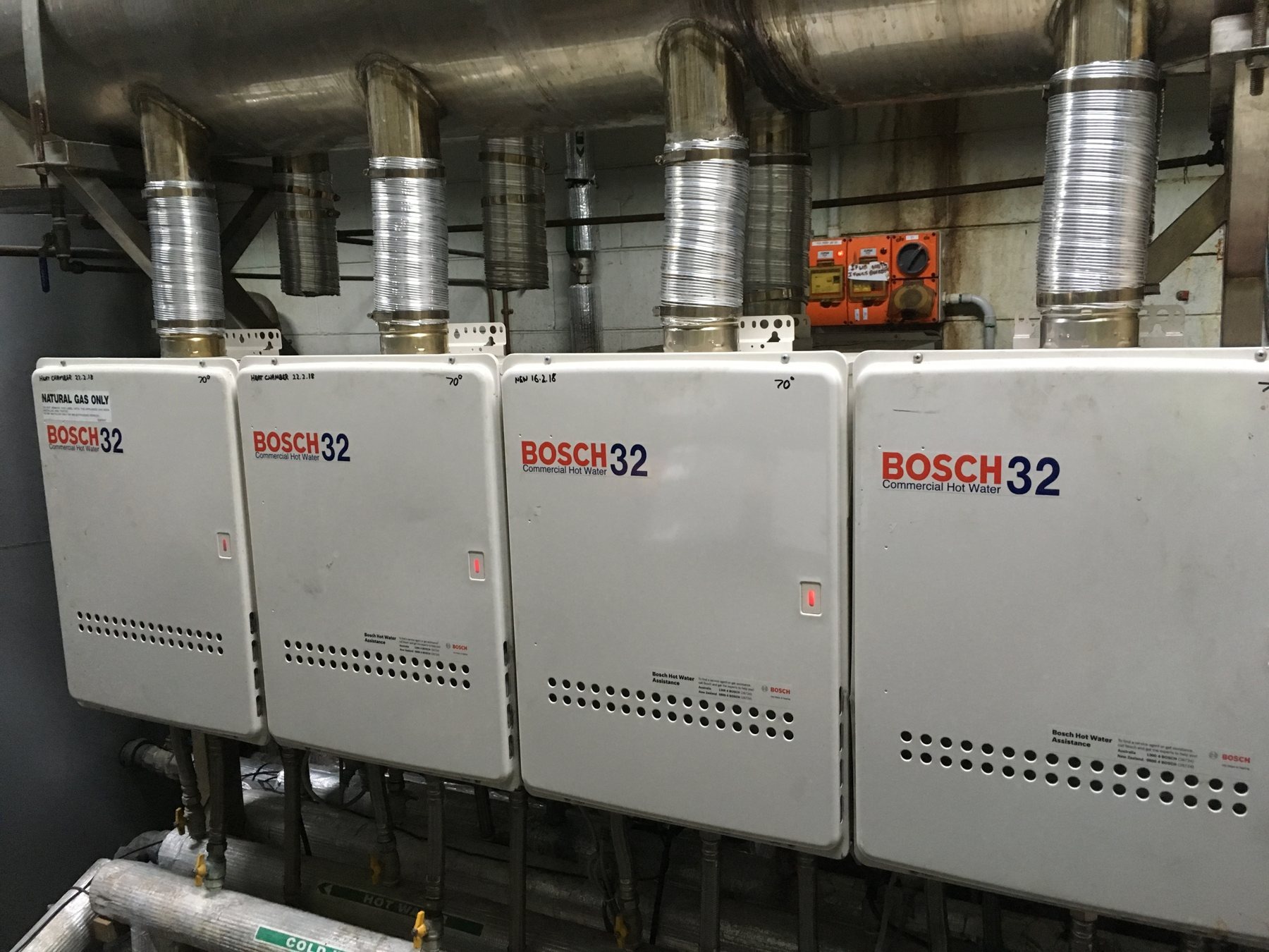 Four-unit commercial water heater bank with a common stainless steel flue header.